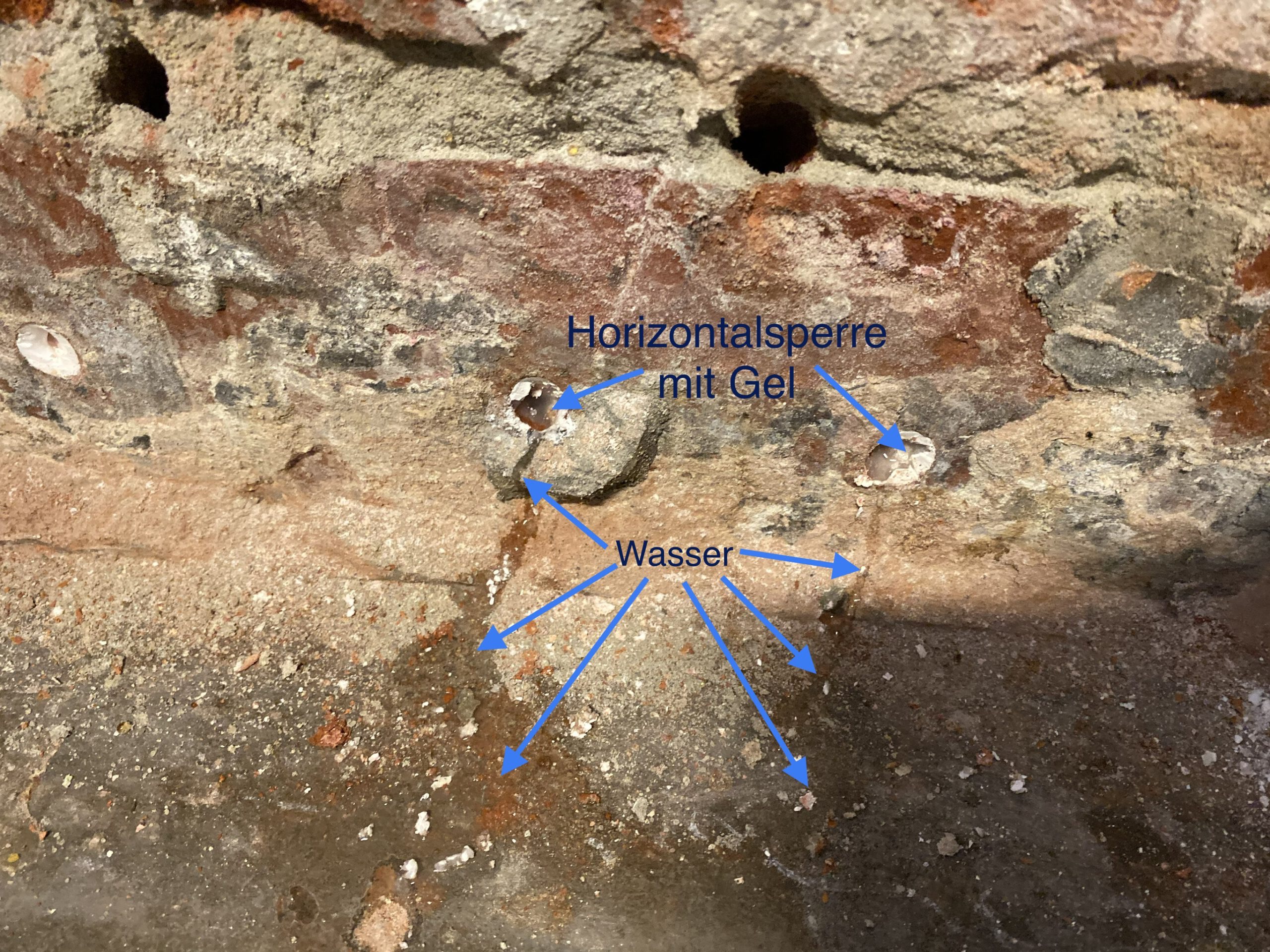 Mauerwerk mit Bohrlöchern und Gel-Injektion, während Wasser trotz Horizontalsperre weiter durch die Wand läuft.